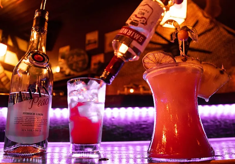 Zwei Cocktails an der Bar, in einen wird gerade Sirup gegossen, das Licht ist lila
