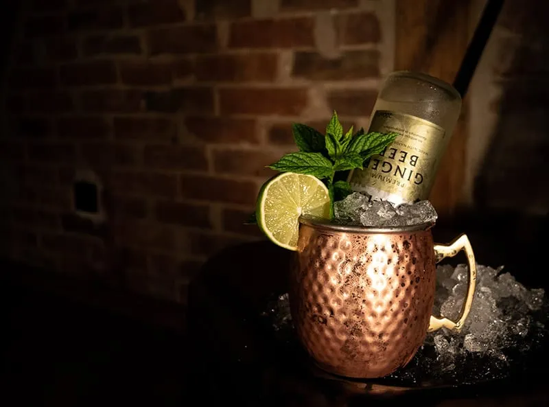 Moscow Mule im Kupferbecher mit Limette und Minze, dahinter eine Flasche Ginger Beer